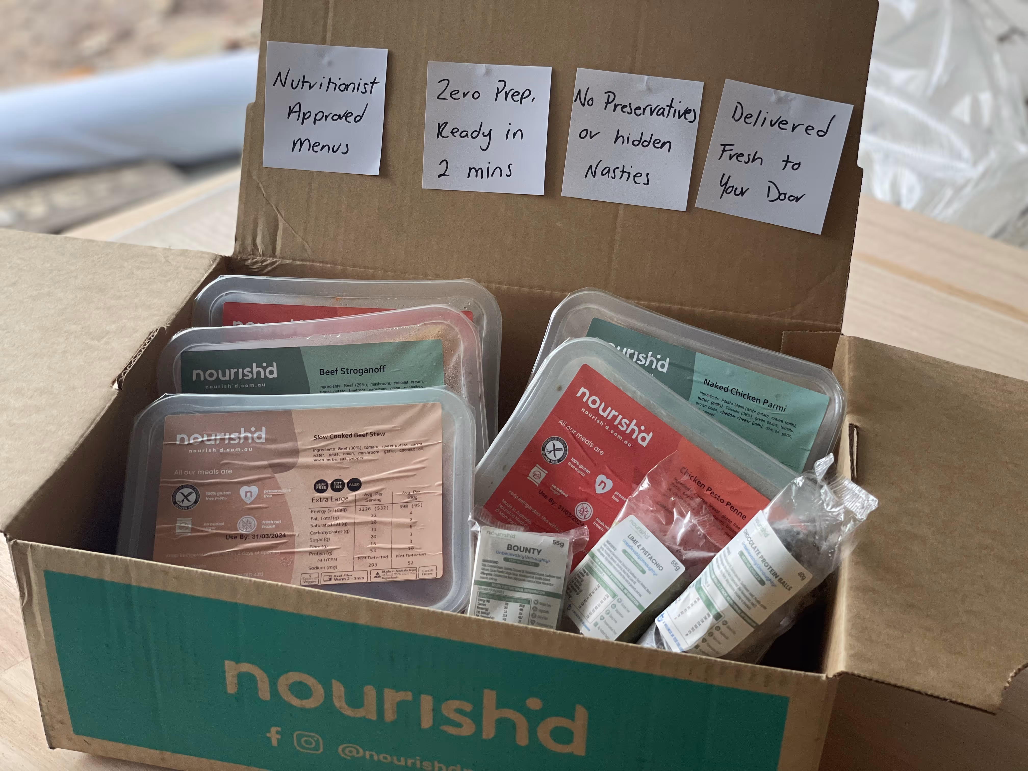 Open Nourishd delivery box containing ready meals and snacks with note cards on the lid reading Nutritionist Approved Menu, Zero Prep Ready in 2 mins, No Preservatives or hidden Nasties, Delivered Fresh to Your Door.
