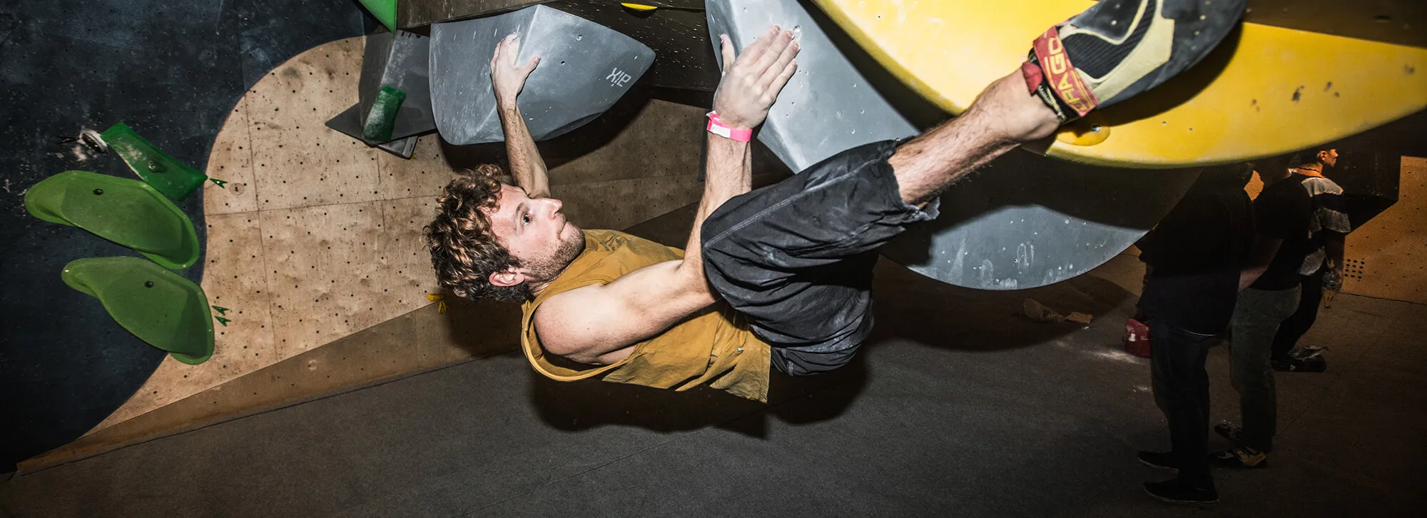 Homme vêtu d'un t-shirt jaune sans manches et d'un pantalon noir escaladant un mur d'escalade intérieur, agrippé à de grandes prises grises et jaunes.