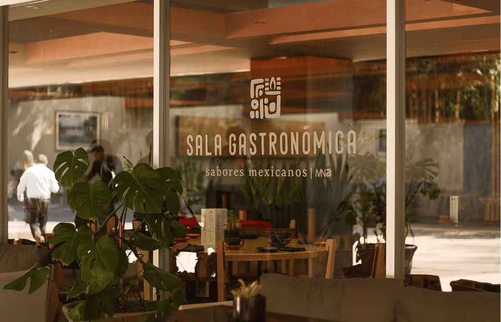 Glass window of a restaurant with the text 'Sala Gastronómica sabores mexicanos' and a leafy green plant in the foreground.