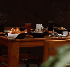 Table set for breakfast with pastries, coffee, juice, and plates of food in a dimly lit dining room.