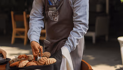 Person wearing a gray apron and blue shirt slicing assorted bread on a wooden board.