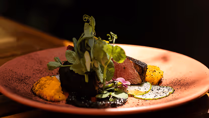 Gourmet dish with seared meat, colorful purees, sliced cucumber, microgreens, and edible flowers on a pink plate.