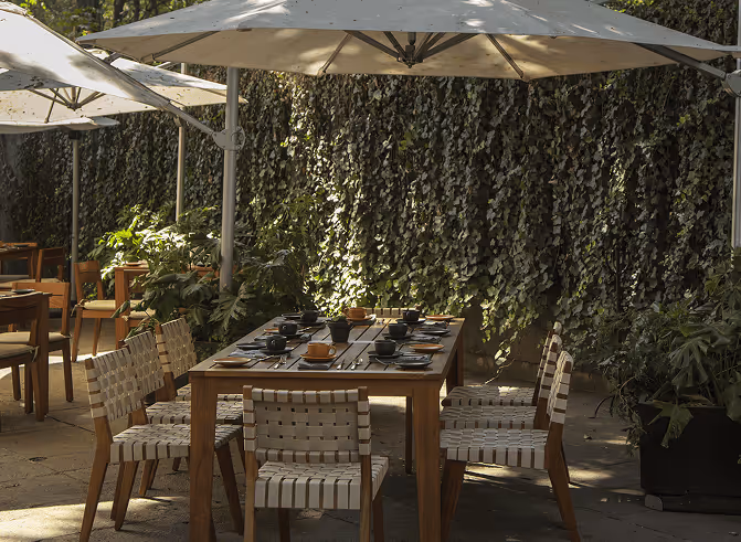 Outdoor dining area with wooden tables and woven chairs under large umbrellas, surrounded by greenery and ivy-covered walls.