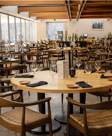 Modern restaurant dining area with wooden tables set with wine glasses, napkins, and menus under a high wooden beam ceiling.