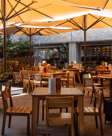 Outdoor restaurant seating area with wooden tables and chairs under large beige umbrellas.