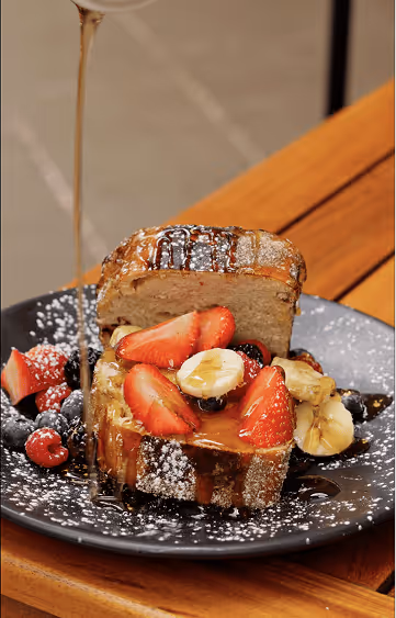 Sliced bread topped with strawberries and bananas on a black plate dusted with powdered sugar and drizzled with syrup.