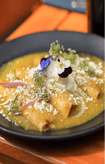 Plate of enchiladas topped with white cheese, cream, and edible flowers, served on a yellow sauce in a dark bowl.