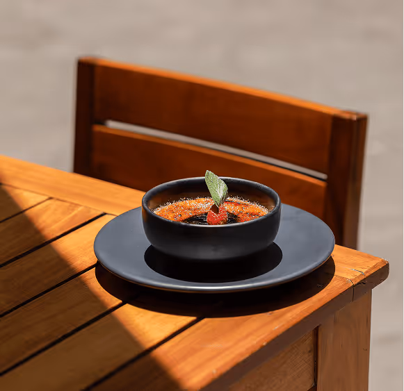Black bowl with crème brûlée garnished with a strawberry and mint leaf on a black plate on a wooden table with a wooden chair in the background.