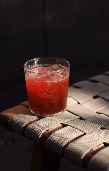 Glass of red iced beverage placed on a woven strap chair against a dark background.