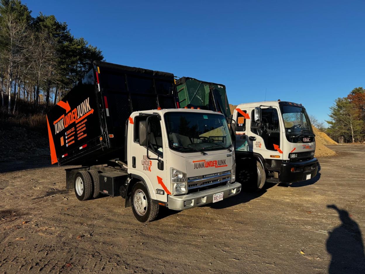 Two white Junk Under Junk trucks with black and orange logo parked on a dirt surface under a clear blue sky.