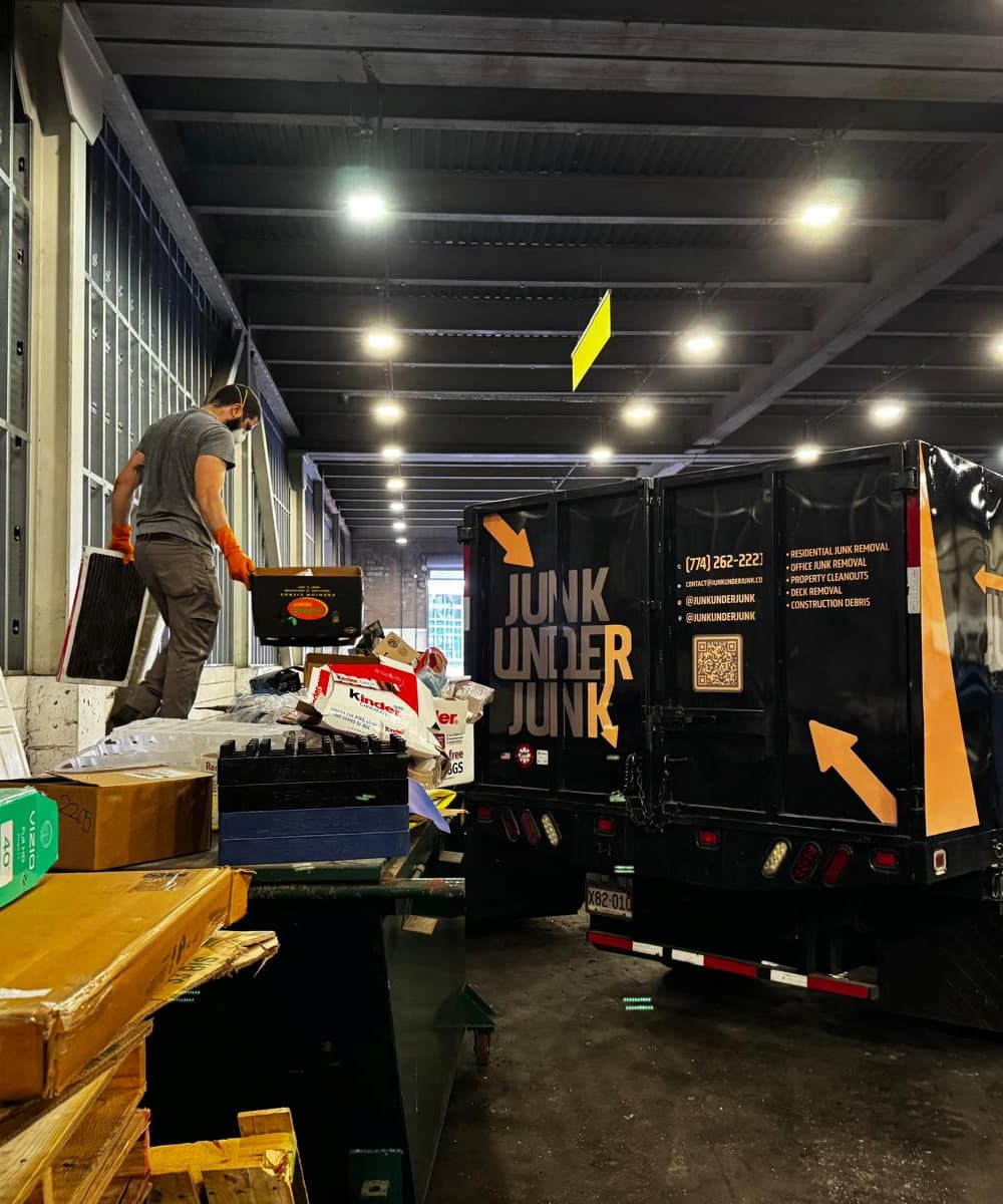 Man wearing orange gloves and a face mask handling junk near a black truck with 'Junk Under Junk' branding inside a warehouse.