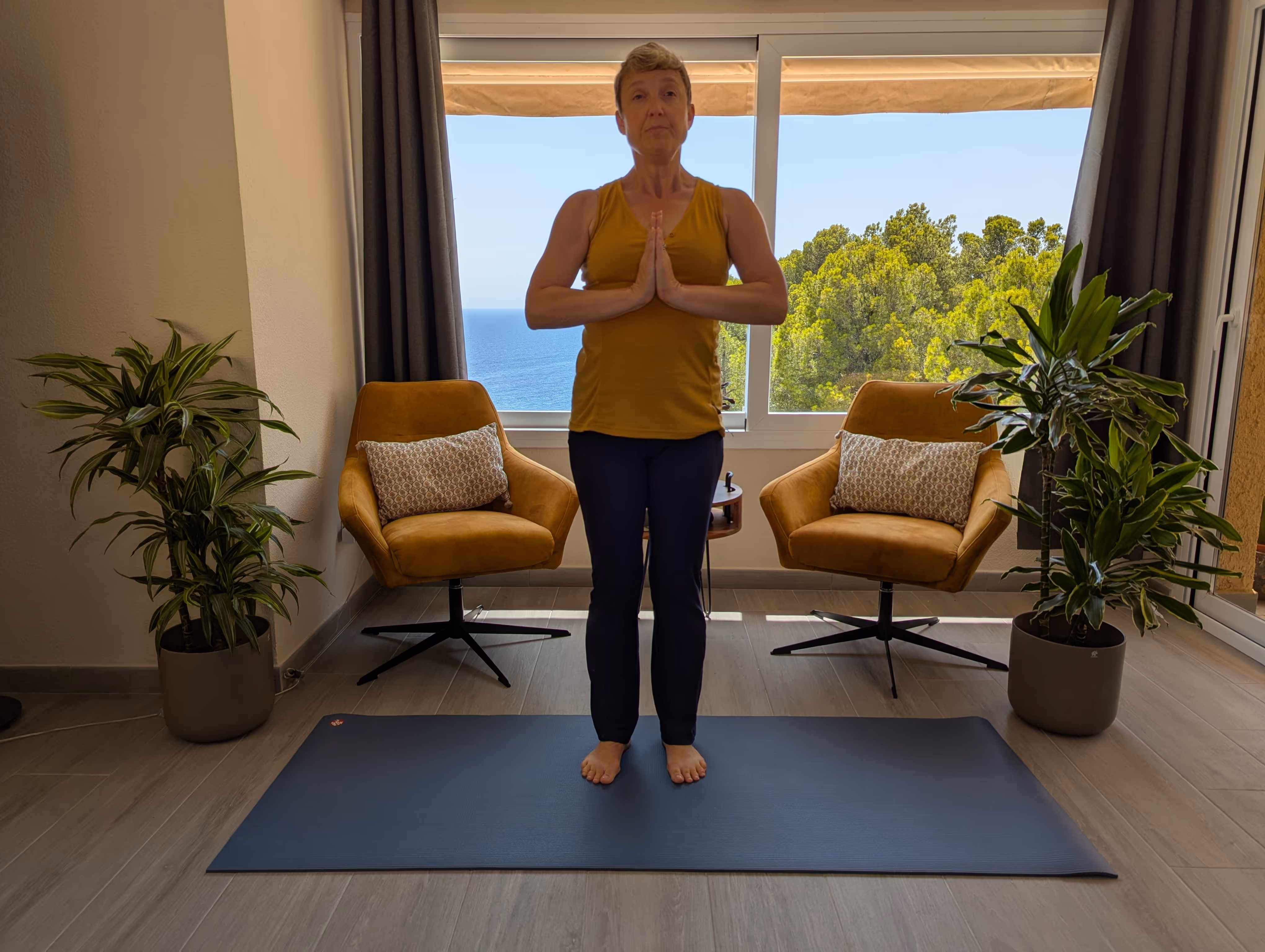 Woman standing in prayer pose on a yoga mat between two chairs, with ocean and trees visible through the window behind her.