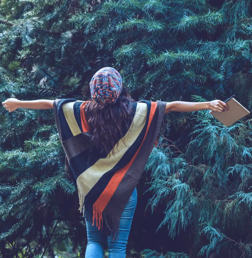 Person standing outdoors with arms wide open, facing lush green trees while holding a notebook.