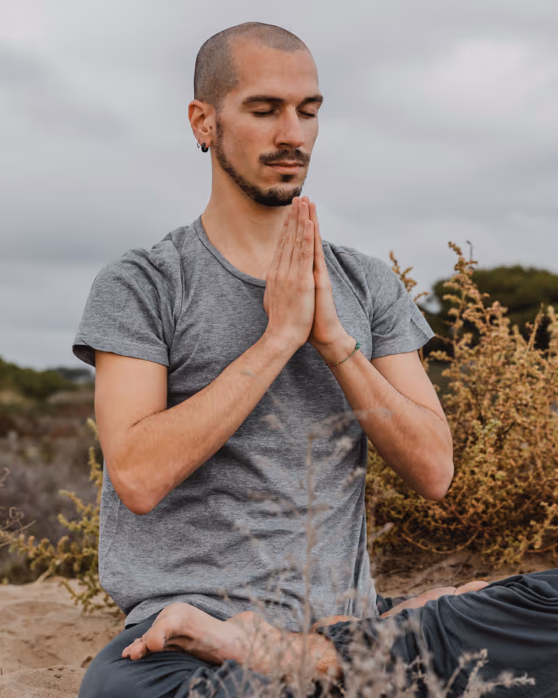 A man sitting outdoors in a meditative pose with eyes closed and hands pressed together in prayer.