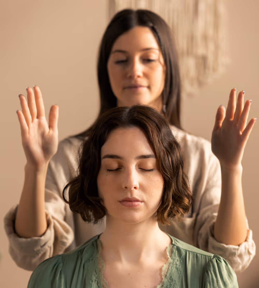 A Reiki practitioner holding her hands above a seated woman’s head as the woman meditates with eyes closed.