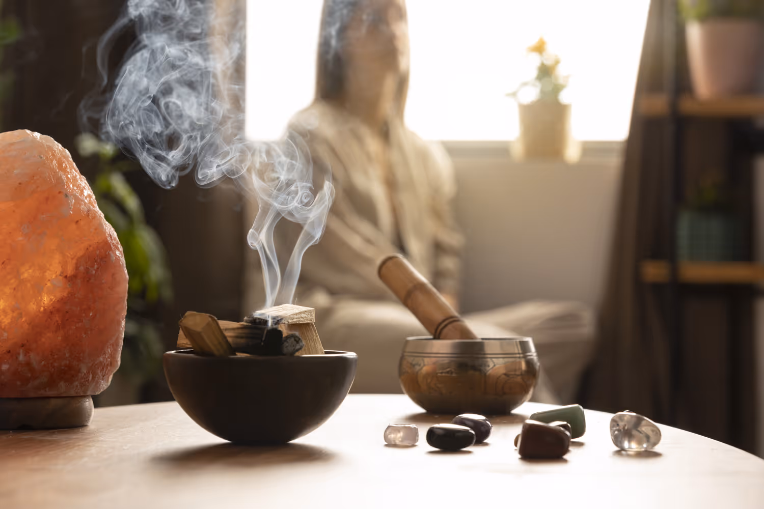 A bowl of burning palo santo with rising smoke on a table beside crystals and a salt lamp, with a person meditating in the blurred background.