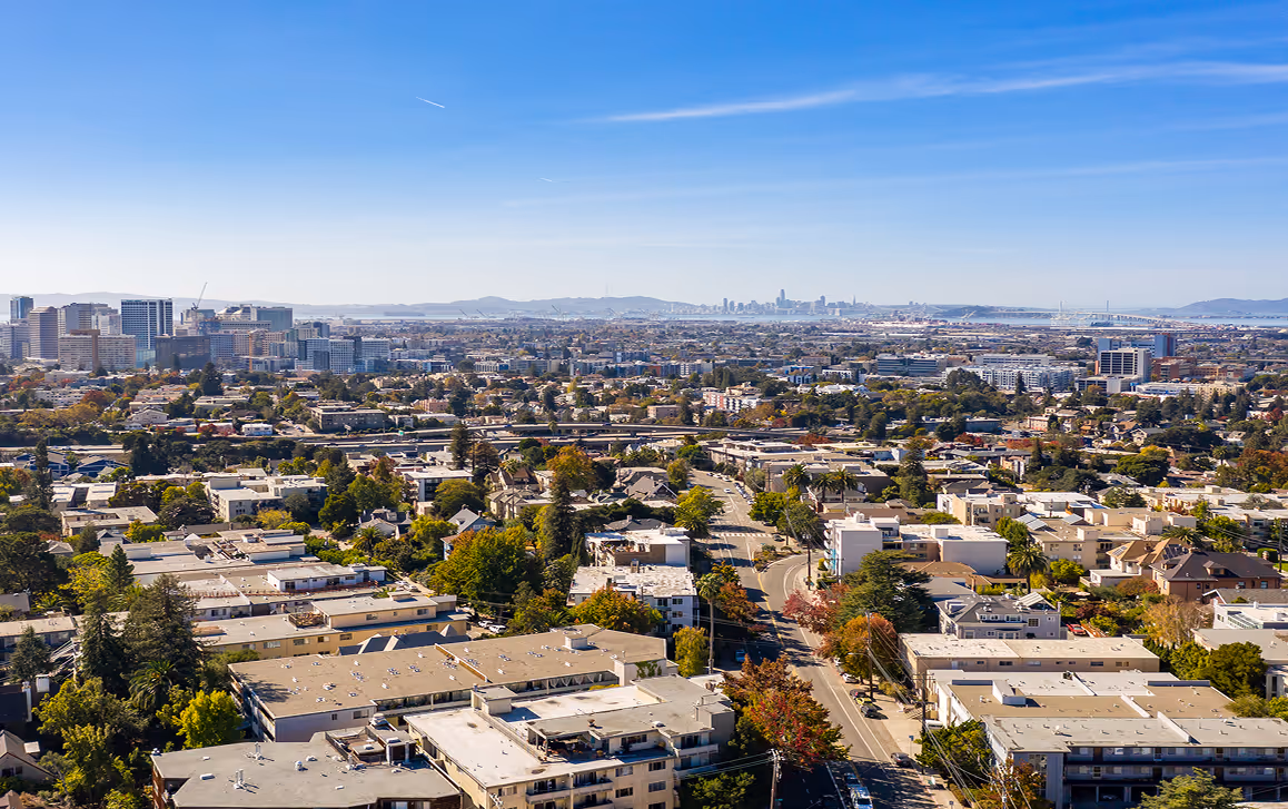 Aerial view of East Bay with residential buildings in the foreground, commercial buildings in the midground, and a downtown skyline with a bridge in the background under a clear blue sky.