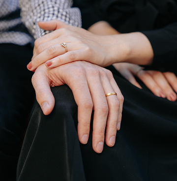 Two hands resting on a black fabric, one wearing a gold wedding band and the other with an engagement ring with a central diamond.