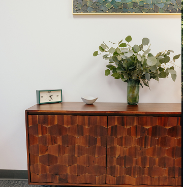 Interior photo of the Arbor Therapy office. Wooden cabinet with hexagonal pattern on doors, topped with a green vase of leafy branches, a clock, and a small bowl against a white wall.