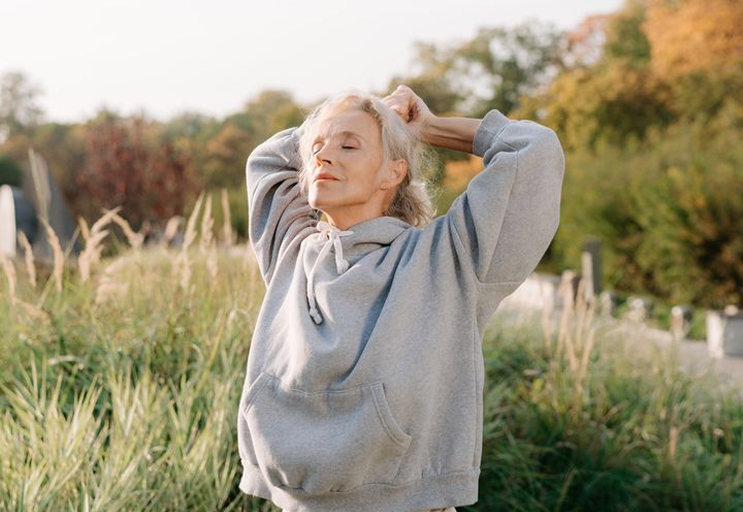 Older woman in a gray hoodie stretching with eyes closed outdoors in a grassy area during autumn.