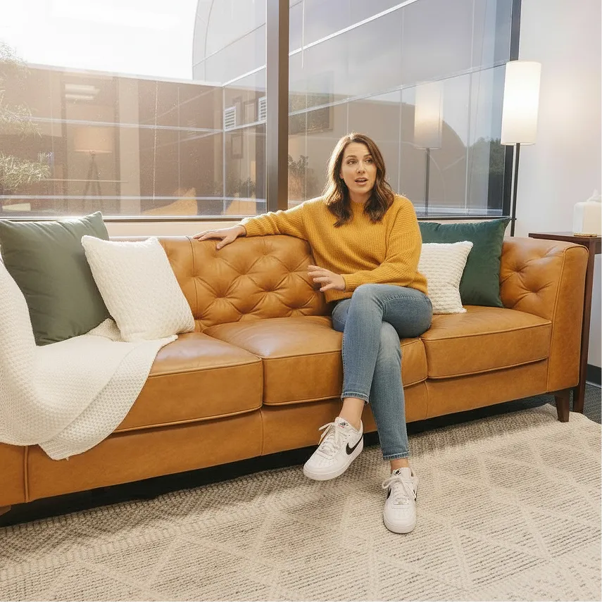A client of Arbor Therapy is in a mustard sweater and jeans sitting on a tan leather couch in a bright modern room with large windows.