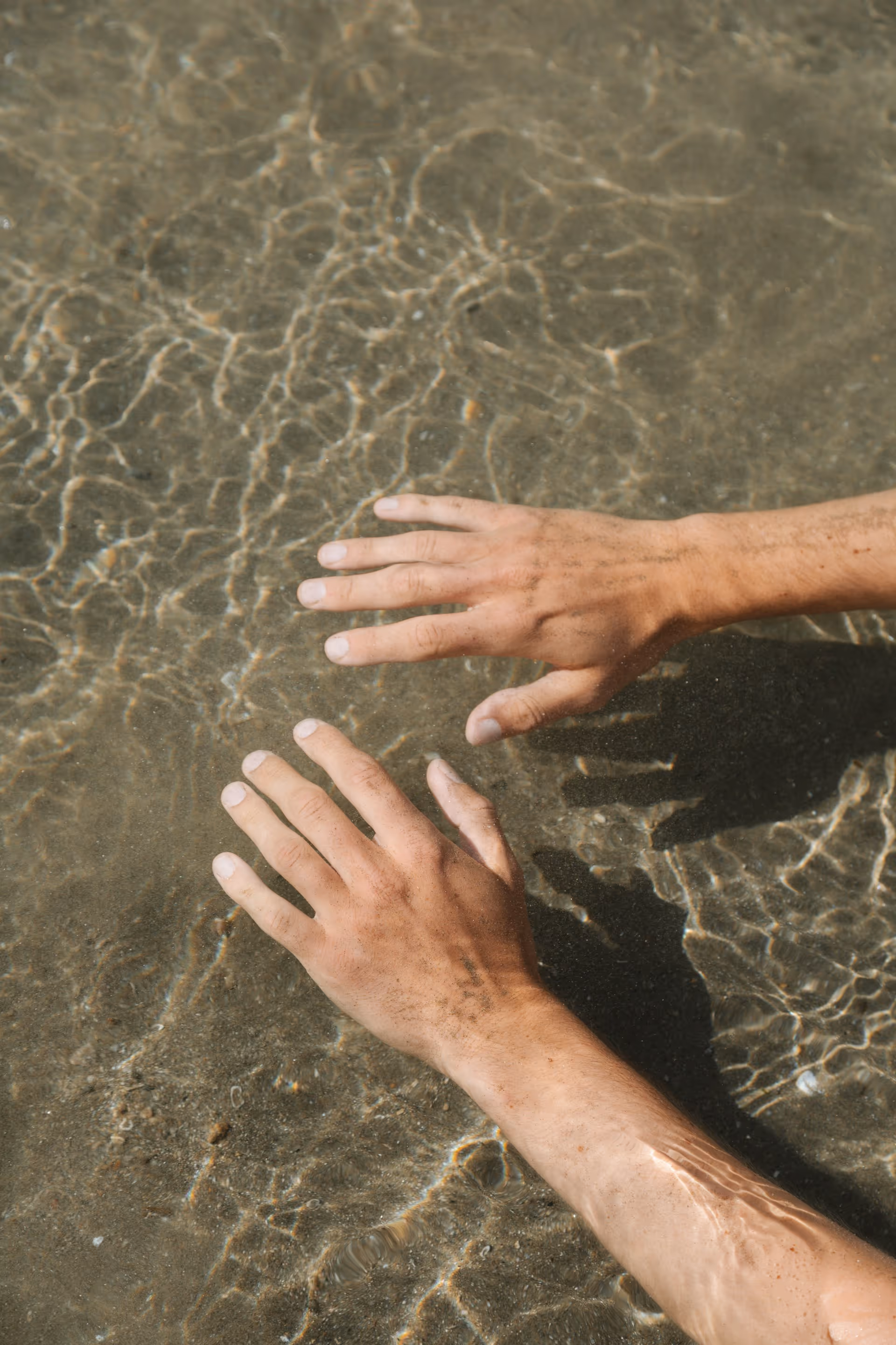 Two hands submerged in clear shallow water with sunlight reflecting on the skin and sandy bottom.