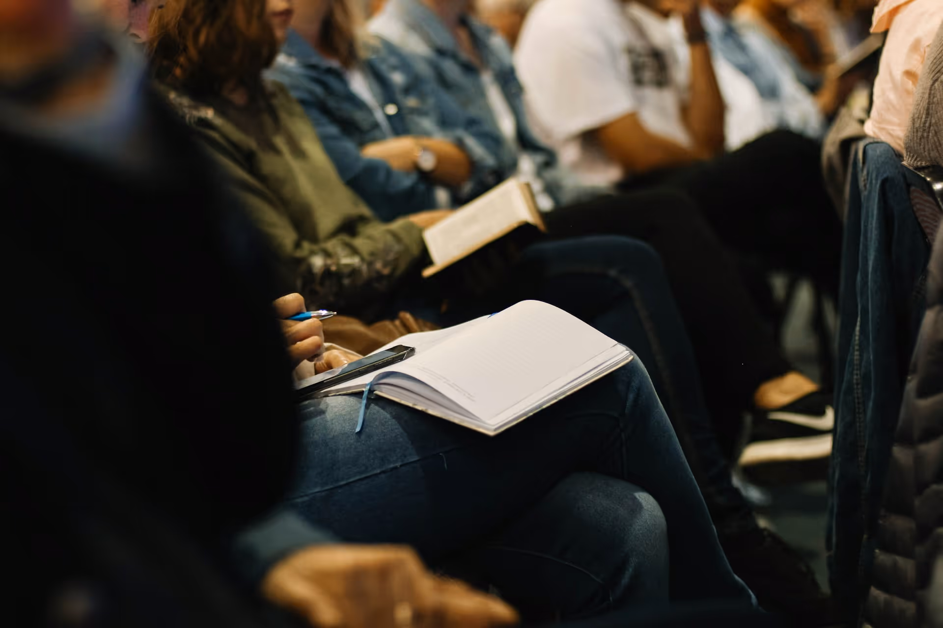 People sitting in a row with notebooks and a book, some taking notes during a meeting or class.