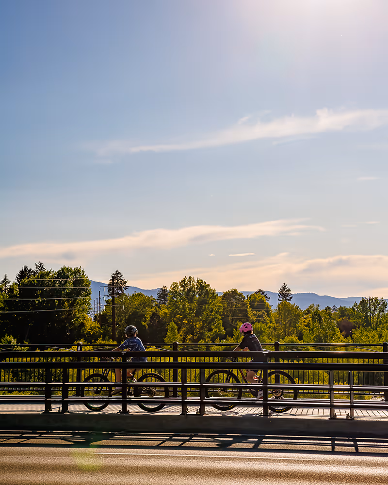 Two cyclists wearing helmets riding on a bridge with trees and mountains in the background under a clear sky.