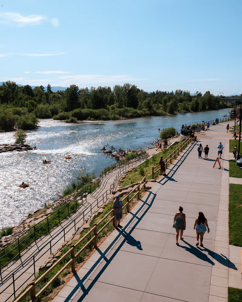 People walking and kayaking near a sunlit river with green trees along the banks on a clear day.