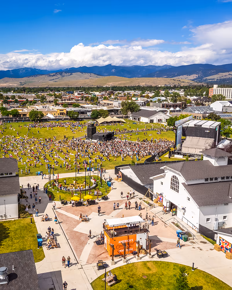 A lively outdoor event with a crowd gathered on a grassy field near stages and buildings, backed by mountains under a partly cloudy sky.