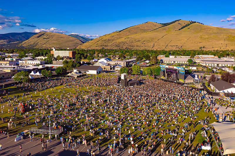 Large outdoor concert with a crowd gathered on green grass near a stage, set against a backdrop of rolling hills and blue sky.