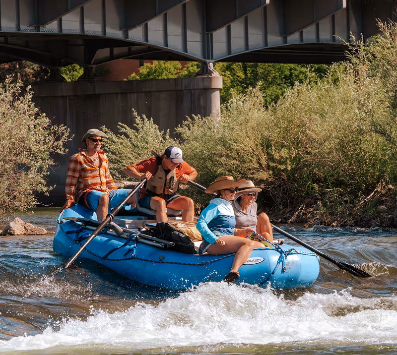 Four people in hats and casual clothing rowing a blue inflatable raft on a river under a bridge surrounded by greenery.