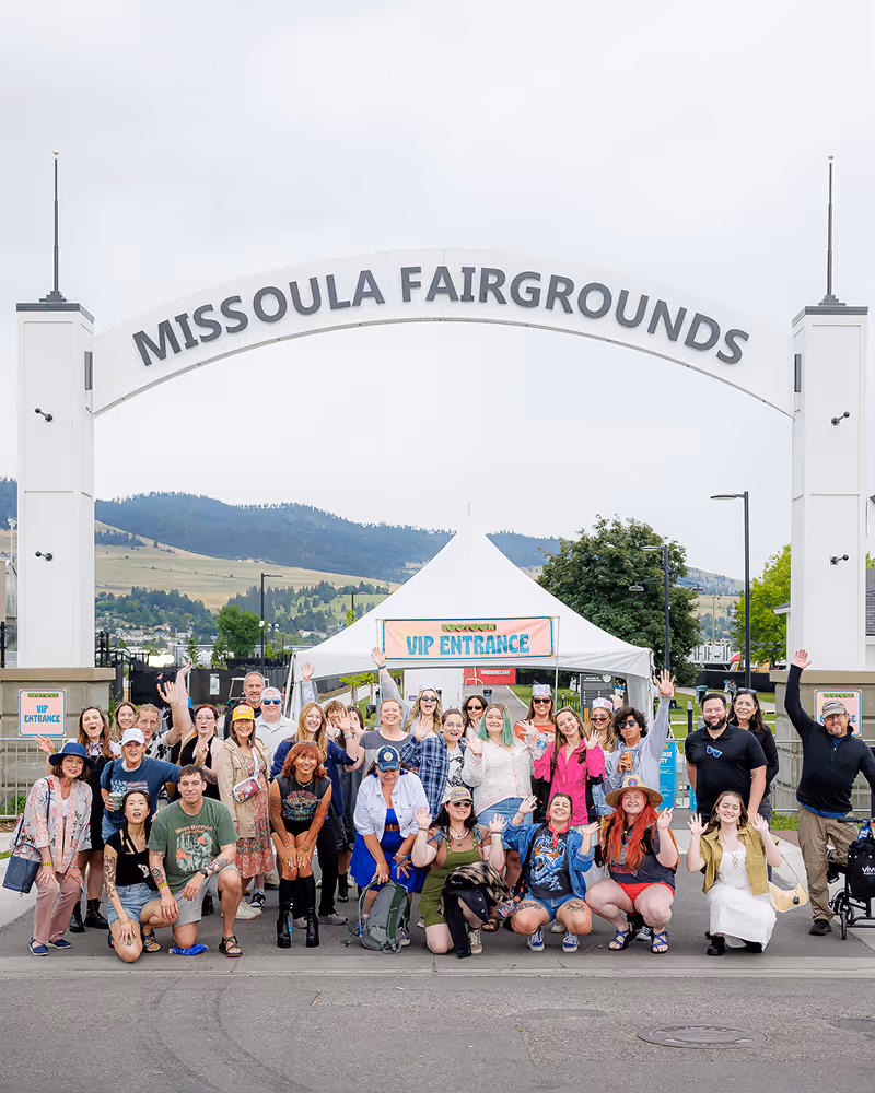 Large diverse group of happy people posing and waving under the Missoula Fairgrounds archway in front of a VIP entrance tent.