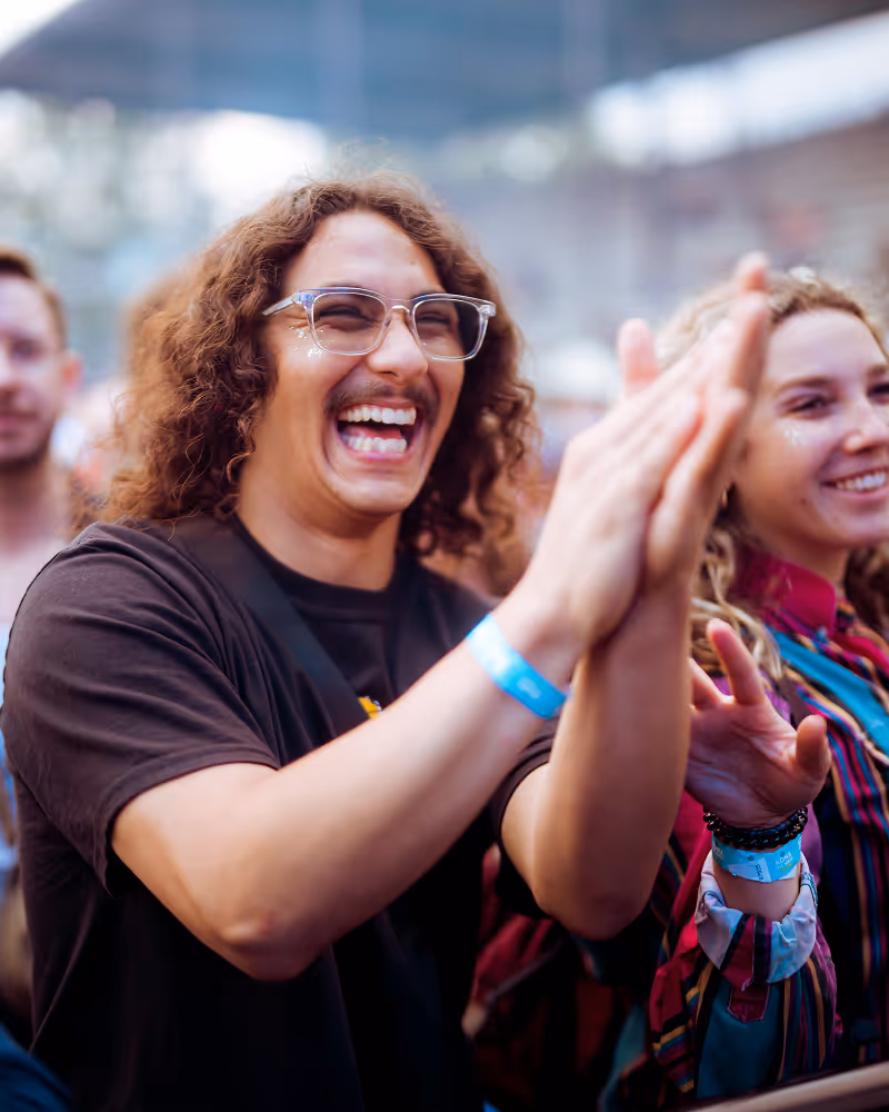 Young man with glasses and curly hair smiling and clapping at an outdoor event, with a woman smiling beside him.