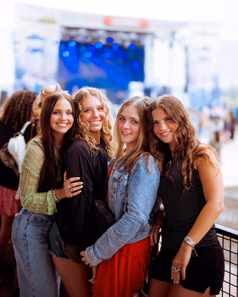 Four young women smiling and posing closely together at an outdoor event with a blurry stage and crowd in the background.