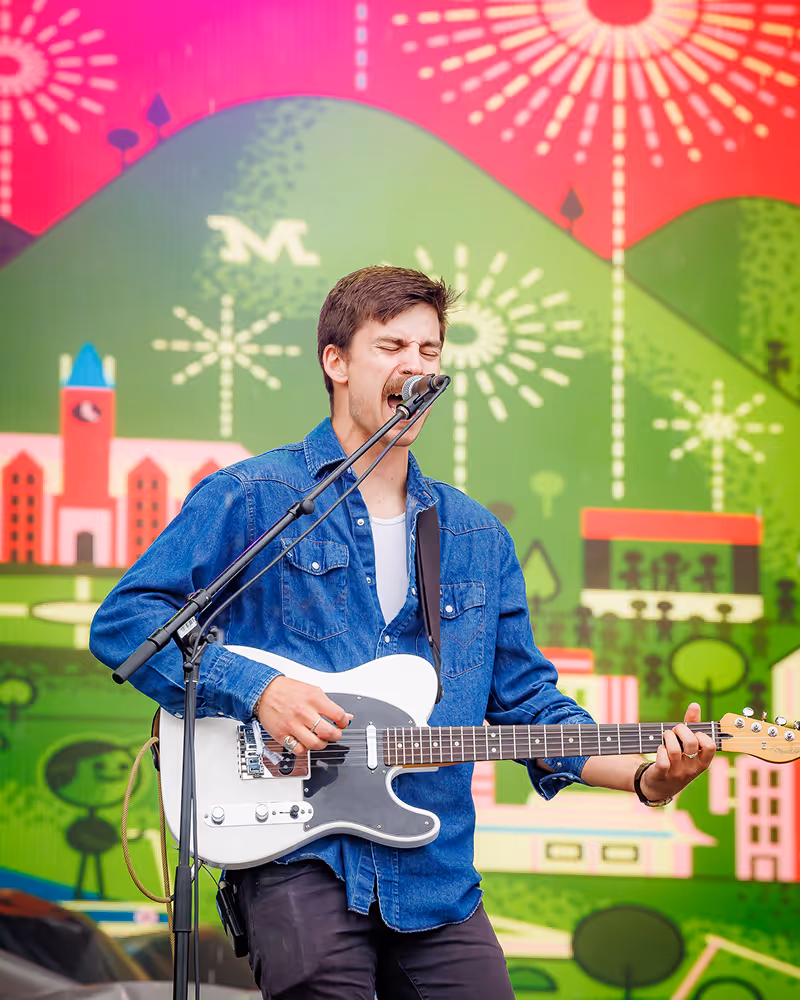 Male musician passionately singing and playing white electric guitar in front of colorful mural backdrop.