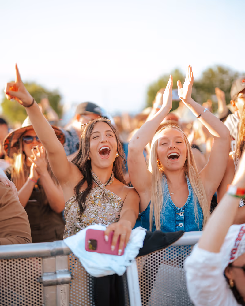 Two young women cheering and raising their hands at an outdoor concert with a crowd in the background.