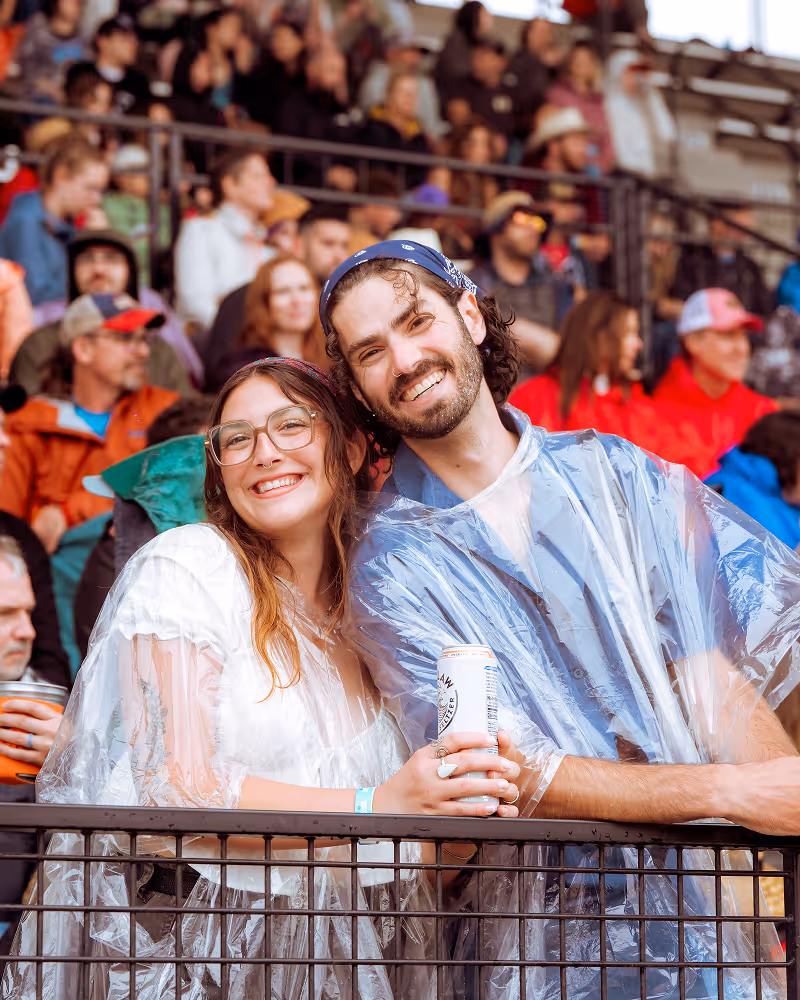 Smiling couple wearing clear rain ponchos at an outdoor event, with a crowd behind them.