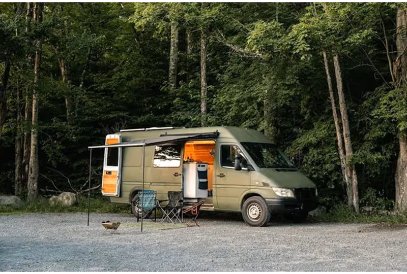 Green camper van with open side door and awning set up in a wooded campsite with two camping chairs and a fire pit.