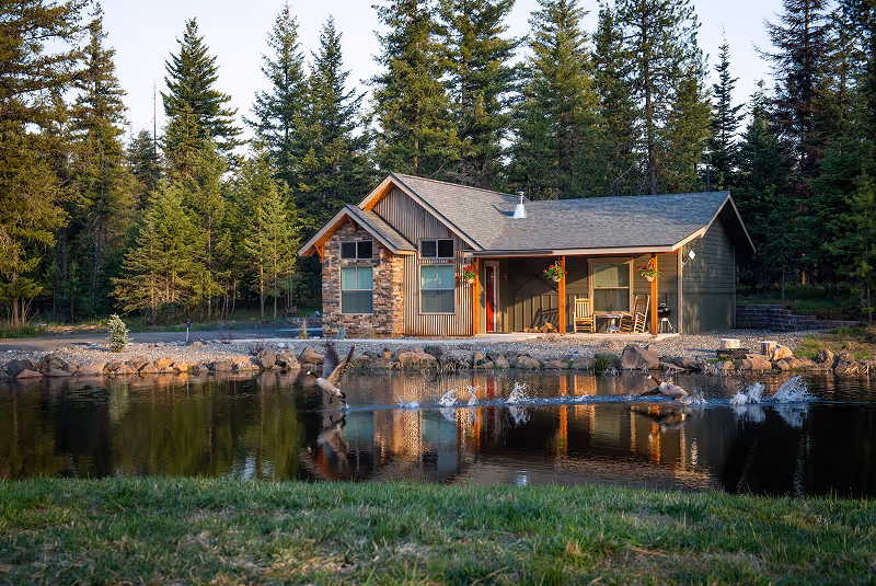 A lakeside cabin with wooden rocking chairs on the porch, surrounded by pine trees and reflected in the water.