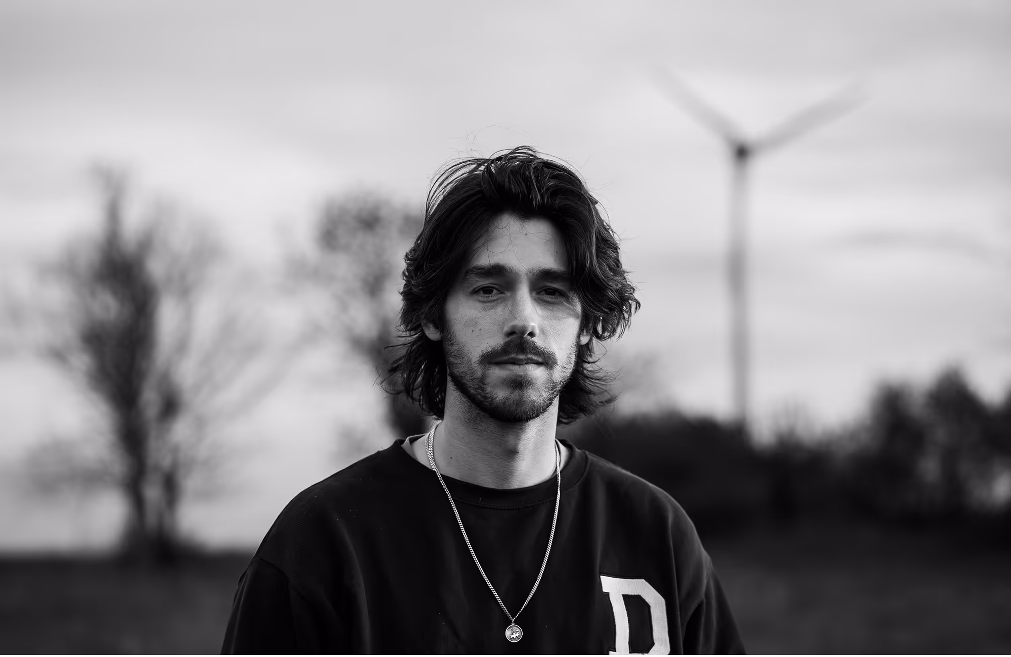 Black and white portrait of a young man with medium-length hair wearing a dark sweatshirt and a pendant necklace outdoors.