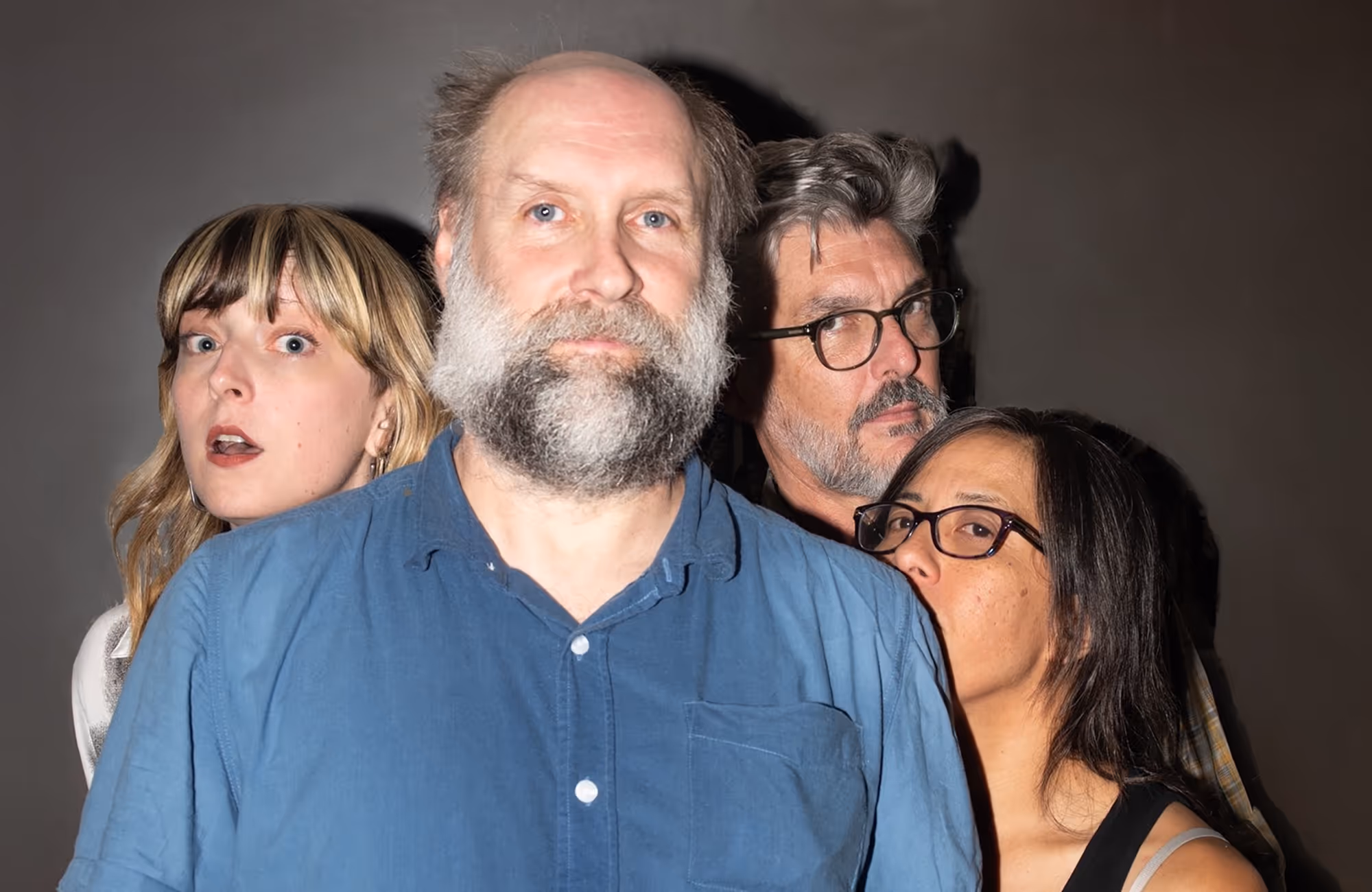 Group of four adults posing closely with neutral expressions against a dark background.