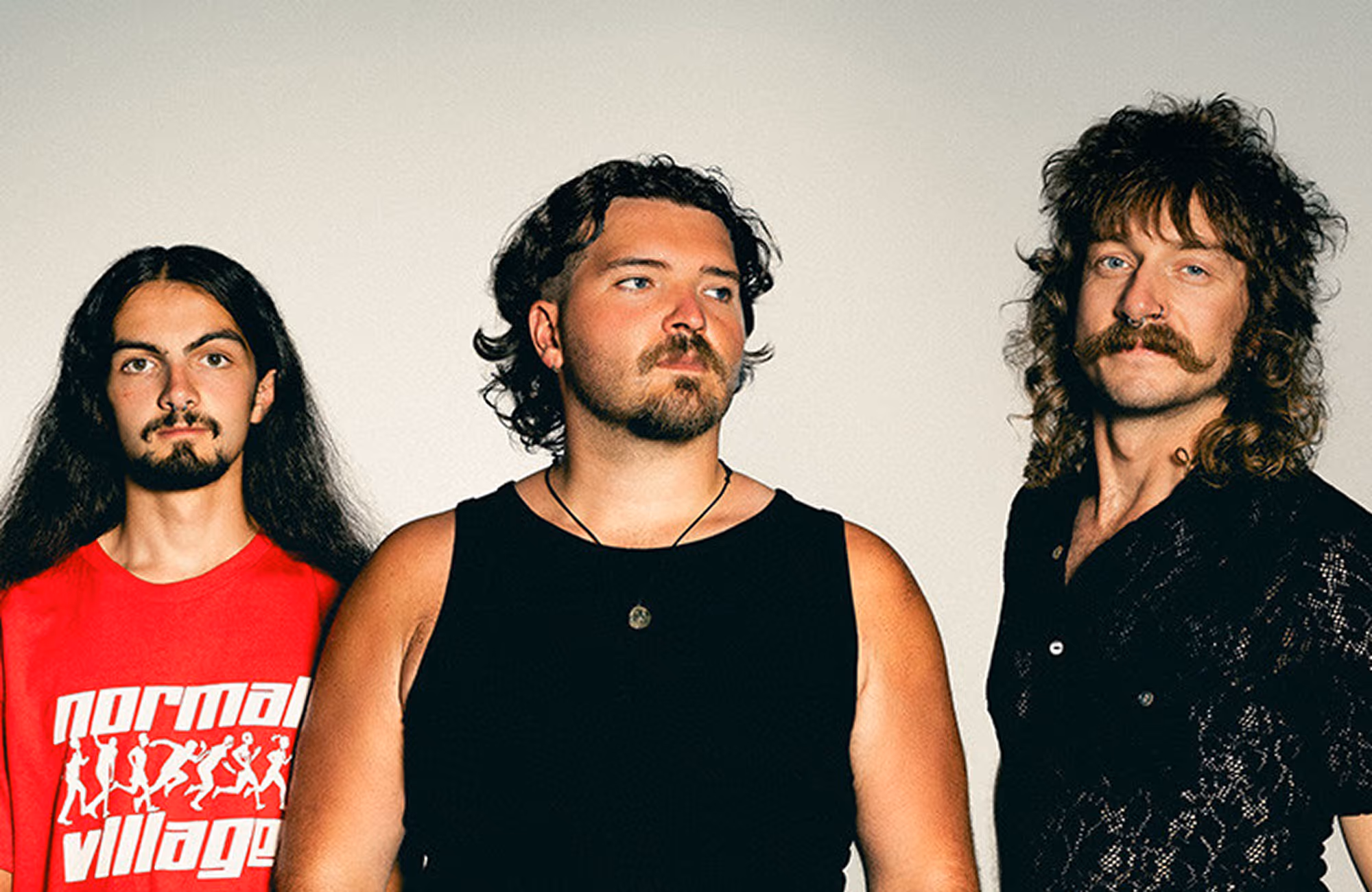 Three men with distinct hairstyles and facial hair standing against a plain background, wearing casual tops.