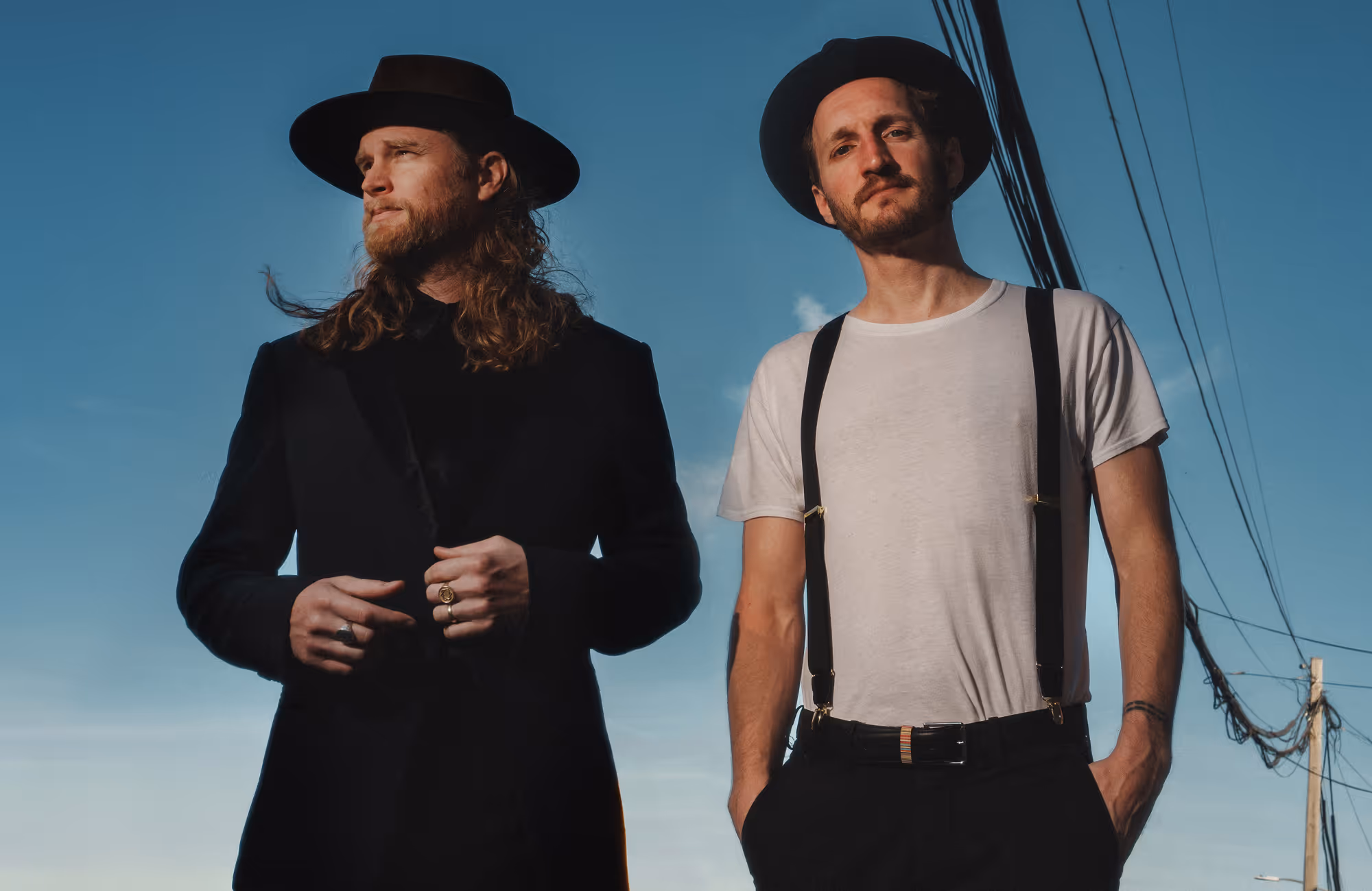 Band promo photo: Lumineers. Two men standing outdoors against a blue sky, one wearing a black hat and coat, the other wearing a black hat, white t-shirt, and suspenders.
