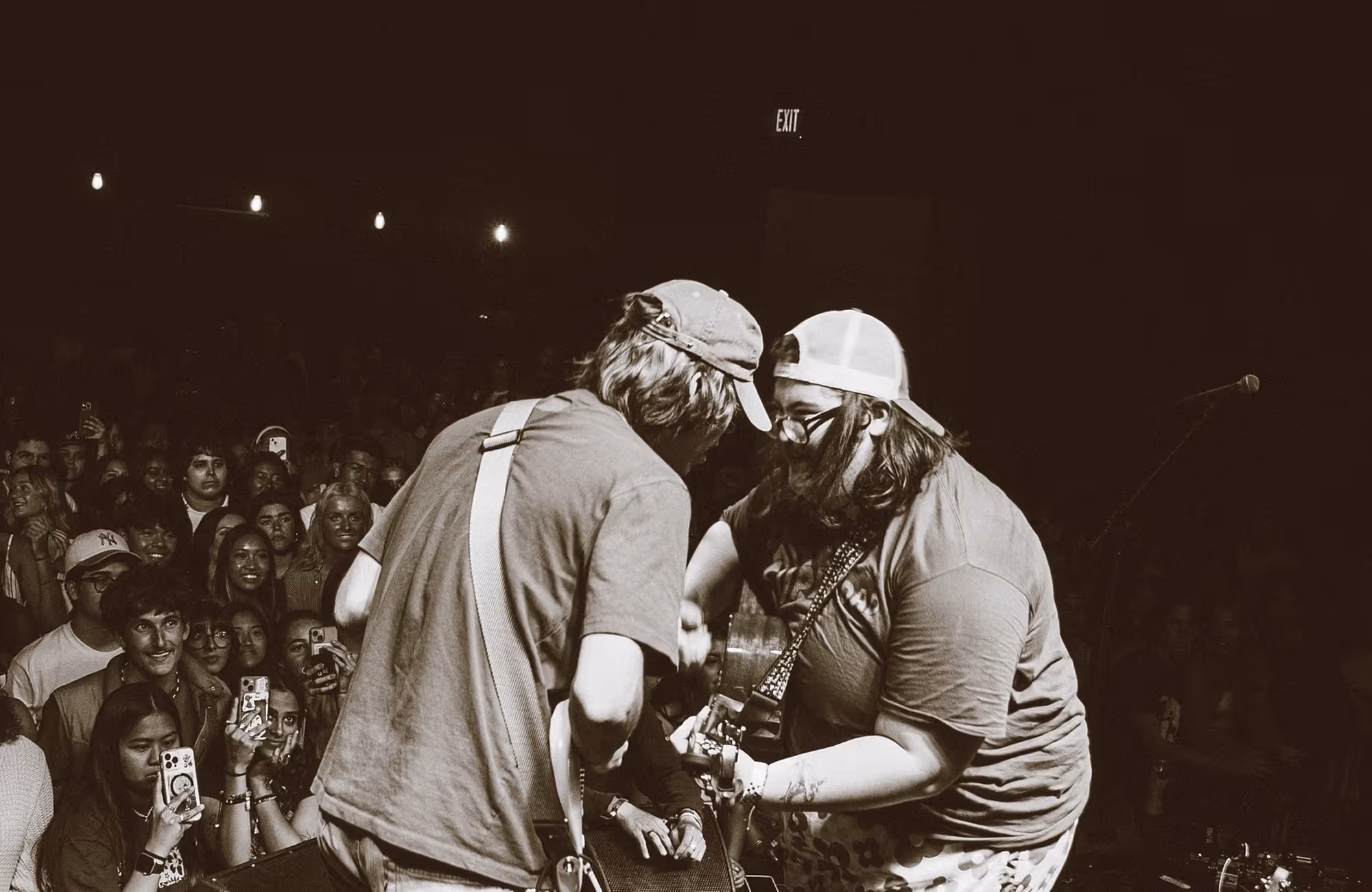 Two musicians play guitars closely together on stage in front of a cheering crowd, with many audience members holding up phones.