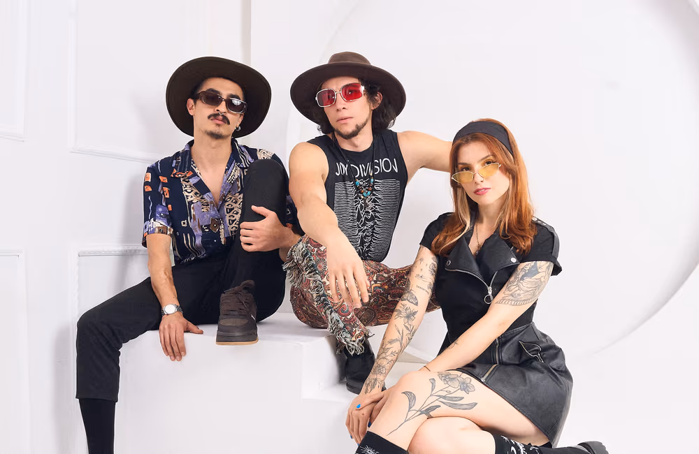 Three young adults in stylish outfits and sunglasses posing against a white background, two men wearing hats and patterned clothes, and a tattooed woman in a black dress.
