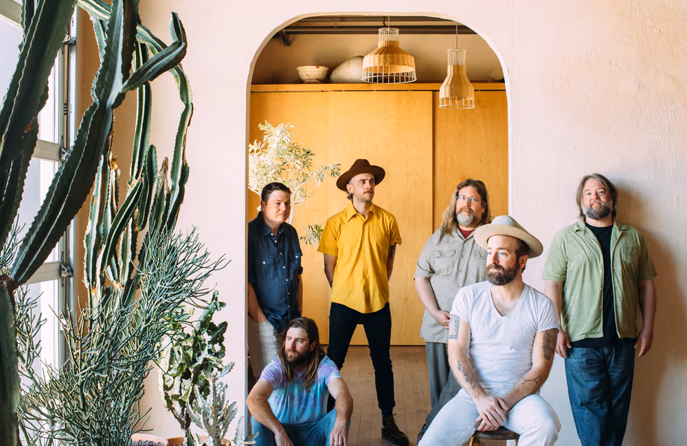 Band promo photo: Trampled by Turtles. Group of six men, two wearing hats, posing in a sunlit room with large cactus plants and wooden lamps overhead.