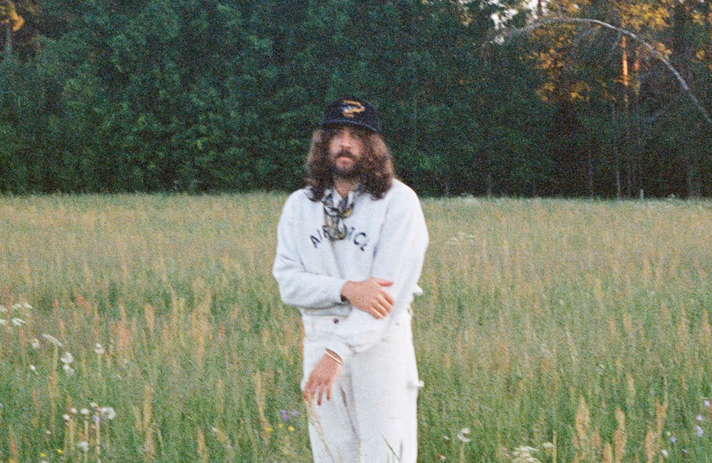 Man with long hair and beard wearing a white outfit and black cap standing in a grassy field with trees in the background.