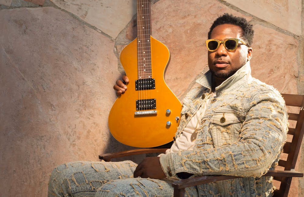 Man wearing textured denim jacket and pants seated on a wooden chair holding a yellow electric guitar, against a stone wall background.