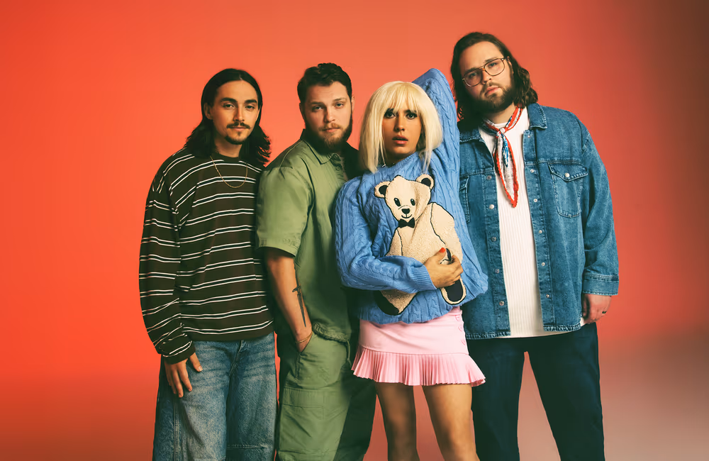 Band promo photo: Rainbow Kitten Surpsie. Four young adults posing against a red background, featuring three men and one woman who is holding a teddy bear pillow.